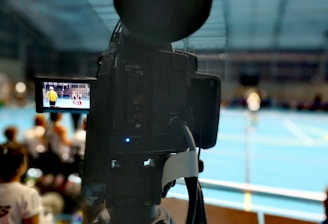 A camera on a tripod is prominently displayed with its viewfinder screen showing footage of an indoor sports event. The background is an indoor sports hall with a blue floor, and numerous spectators are faintly visible.