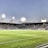 A large soccer stadium filled with spectators under bright floodlights. The field is green and well-maintained with players actively engaged in a match. Advertising banners line the perimeter of the stadium. A cameraman is positioned near the foreground, capturing the action on the field. The sky above is clear with a hint of dusk.