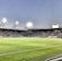 A large soccer stadium filled with spectators under bright floodlights. The field is green and well-maintained with players actively engaged in a match. Advertising banners line the perimeter of the stadium. A cameraman is positioned near the foreground, capturing the action on the field. The sky above is clear with a hint of dusk.