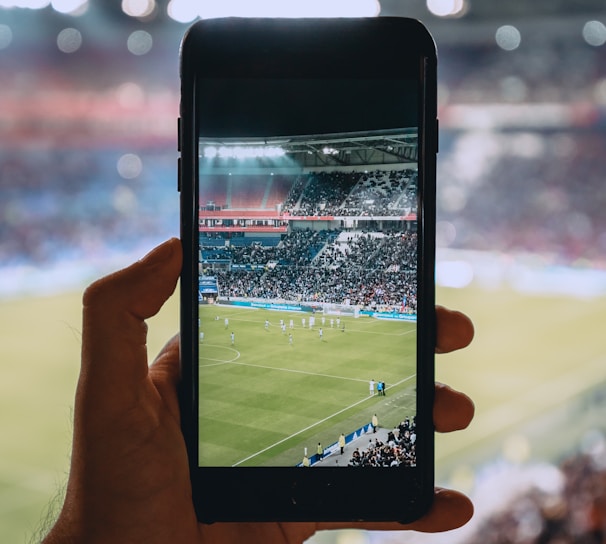 A hand holding a smartphone captures a live view of a soccer stadium filled with fans and players on the field. The image on the smartphone screen is crisp, showing a wide-angle view of the game in action.