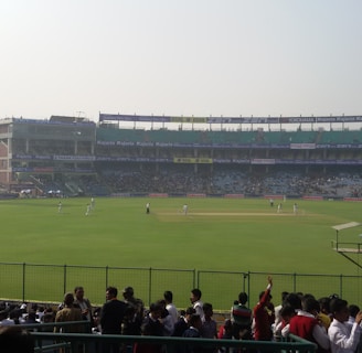 A cricket stadium filled with spectators, with players on the field wearing white uniforms. The stands are populated, and large floodlights are visible. Advertisements are displayed around the stadium, and a bright digital screen is on the left side.
