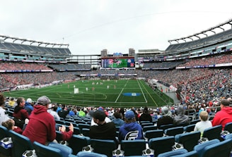 A large sports stadium filled with spectators. The field is green with white markings, and players in red and white uniforms are actively engaged in a game. The stands are packed with people wearing various colored clothing, and some sections of the crowd are more densely populated. Digital screens and advertisements are visible around the stadium, adding to the vibrant atmosphere.