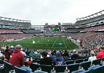 A large sports stadium filled with spectators. The field is green with white markings, and players in red and white uniforms are actively engaged in a game. The stands are packed with people wearing various colored clothing, and some sections of the crowd are more densely populated. Digital screens and advertisements are visible around the stadium, adding to the vibrant atmosphere.