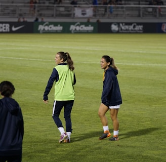 Two people are on a soccer field, wearing sports attire. One person is wearing a green vest, and the other is in a blue jacket and white shorts. The setting appears to be during a sports event, with adverts in the background and a partially filled stadium.