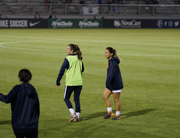 Two people are on a soccer field, wearing sports attire. One person is wearing a green vest, and the other is in a blue jacket and white shorts. The setting appears to be during a sports event, with adverts in the background and a partially filled stadium.