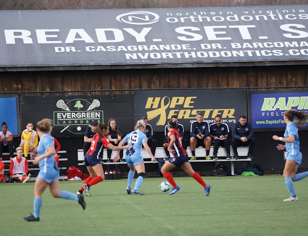 Players engage in a competitive soccer game on a field while others are seated on the bench watching attentively. The action is taking place in front of a wooden structure displaying multiple advertisements, including orthodontics, lacrosse, and field hockey.