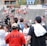 A lively gathering in an outdoor sports venue with a group of people, some wearing matching shirts labeled 'National Champions.' Several individuals are holding cameras and microphones, indicating a media presence. The background shows blurred spectators sitting on bleachers.