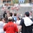 A lively gathering in an outdoor sports venue with a group of people, some wearing matching shirts labeled 'National Champions.' Several individuals are holding cameras and microphones, indicating a media presence. The background shows blurred spectators sitting on bleachers.