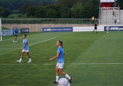 A soccer game scene on a green grass field with players wearing light blue jerseys and white shorts. A goalpost is visible in the background, and there are people sitting or standing on the bleachers. Advertising banners are displayed along the sidelines.