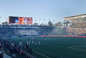 A large sports stadium filled with spectators. The field is occupied by soccer players, some walking and others scattered across the pitch. A massive screen displays a person, and flags fly on top of the stadium roof. The audience is dense, and the atmosphere seems lively with a visible banner showing 'BLUE WHITE & GOLD'.