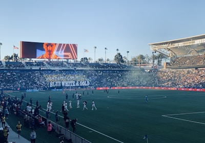 A large sports stadium filled with spectators. The field is occupied by soccer players, some walking and others scattered across the pitch. A massive screen displays a person, and flags fly on top of the stadium roof. The audience is dense, and the atmosphere seems lively with a visible banner showing 'BLUE WHITE & GOLD'.