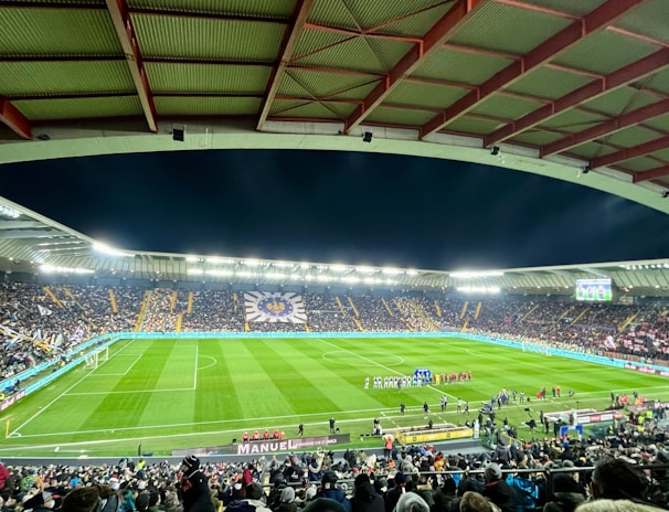 A large football stadium filled with spectators. The field is brightly lit, and two teams are positioned on the pitch. The stadium is covered, with a visible roof structure and stands filled with fans. Scoreboards are visible displaying game information.