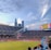 A sports stadium filled with spectators watching a soccer match. Players are on the field in action, and a large scoreboard displays information about the game. The stadium has blue seating and is surrounded by tall city buildings visible in the background against a partly cloudy sky. Fans are dressed casually, some waving flags and wearing team colors.