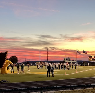 A sports field with a football game unfolding under a vibrant sunset sky, featuring a large inflatable mascot and a scoreboard displaying the score. Players and coaches are visible on the green field, surrounded by a track and some flags waving in the breeze.