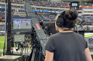 A person with headphones is operating a large camera setup at a sports stadium. The camera equipment is focused on a screen showing a live sports event taking place on a football field. The stadium is filled with spectators, and the atmosphere is busy with technical setup and monitors.
