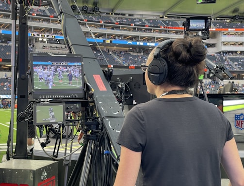 A person with headphones is operating a large camera setup at a sports stadium. The camera equipment is focused on a screen showing a live sports event taking place on a football field. The stadium is filled with spectators, and the atmosphere is busy with technical setup and monitors.