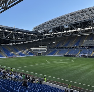 A modern sports stadium with a large, green soccer field and surrounding seating areas. Blue and yellow seats are visible, with some spectators scattered across the stands. The stadium is partially covered with a steel lattice roof, showing a clear blue sky. Several security personnel in high-visibility vests are present.
