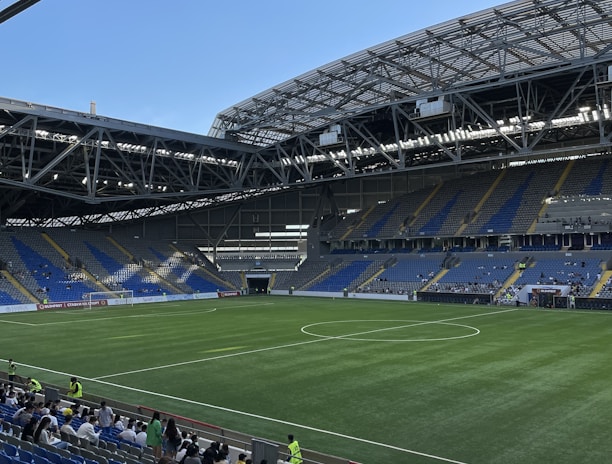 A modern sports stadium with a large, green soccer field and surrounding seating areas. Blue and yellow seats are visible, with some spectators scattered across the stands. The stadium is partially covered with a steel lattice roof, showing a clear blue sky. Several security personnel in high-visibility vests are present.
