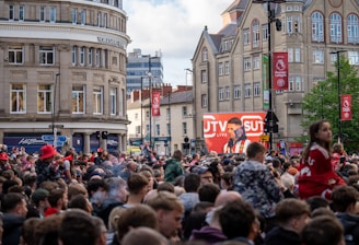 A large crowd of people, many wearing red and white, gather in a city square. They are watching an event broadcast on a large screen, which displays a man speaking into a microphone with logos suggesting a sporting event. Surrounding buildings are historical and commercial, with banners indicating a connection to the Premier League.