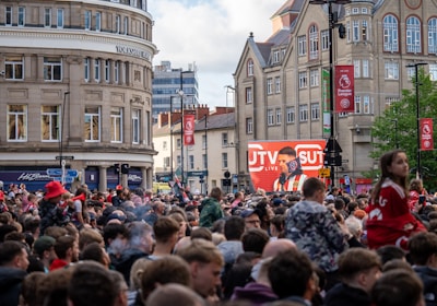 A large crowd of people, many wearing red and white, gather in a city square. They are watching an event broadcast on a large screen, which displays a man speaking into a microphone with logos suggesting a sporting event. Surrounding buildings are historical and commercial, with banners indicating a connection to the Premier League.