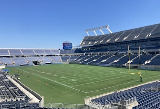 A large, empty sports stadium with blue seats surrounding a well-maintained green grass field. Bright sunlight creates clear shadows, and there are two goal posts on opposite ends of the field. A big electronic scoreboard displays some graphics, and the sky is clear and blue.