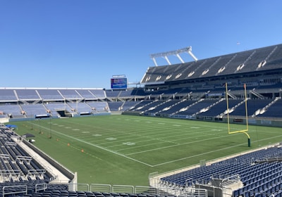 A large, empty sports stadium with blue seats surrounding a well-maintained green grass field. Bright sunlight creates clear shadows, and there are two goal posts on opposite ends of the field. A big electronic scoreboard displays some graphics, and the sky is clear and blue.
