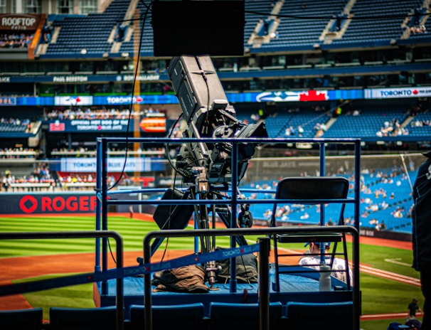 A professional-grade video camera is mounted on a platform overlooking a baseball field. The stadium seats in the background are filled with some spectators, and various advertisements are visible around the field. A staff member wearing a jacket with 'STAFF' written on the back is positioned near the camera.